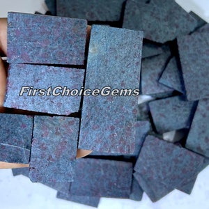 Może przedstawiać: Stosik ciemnoniebieskich i fioletowych odłamków kamieni szlachetnych z czerwonymi plamkami. Odłamki są prostokątne i mają szorstką, naturalną fakturę. Odłamki są ułożone w stos, niektóre z nich zachodzą na siebie. Odłamki są trzymane w dłoni, palce są widoczne. Tekst "FirstChoiceGems" jest widoczny na zdjęciu.