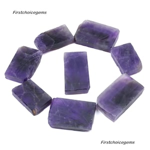 Puede incluir: Una colección de piedras de amatista en bruto, que muestran un tono púrpura intenso con vetas más oscuras y contrastantes. Las piedras tienen forma irregular, con una textura rugosa y natural. La imagen incluye el texto "Firstchoicegems".