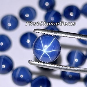 Puede incluir: Primer plano de una piedra preciosa de zafiro estrellado azul con un patrón de estrella de seis puntas. La piedra preciosa está sujeta con pinzas y rodeada de otros zafiros estrellados sueltos.