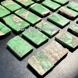 May include: A collection of green and gray gemstone cabochons, cut into rectangular shapes. The stones have a mottled pattern and are arranged in rows on a black surface. The text "FirstChoiceGems" is visible in the image.