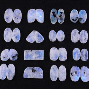 Può includere: Una collezione di pietre preziose di pietra di luna, ognuna con una base bianco latte con inclusioni blu e nere. Le pietre sono tagliate in varie forme, tra cui ovali, rettangoli e triangoli, su sfondo nero.