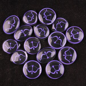 May include: A collection of black, glossy, glass-like stones with a purple bow and arrow design. The stones are in various shapes, including round and oval, and are arranged on a dark surface. The bow and arrow design is centered on each stone.