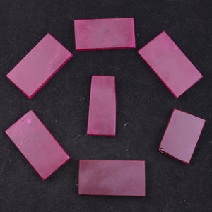 May include: Seven rectangular, polished, ruby-colored gemstones arranged on a black surface. The gemstones vary slightly in size and shape, with a deep, translucent hue. The arrangement suggests a potential for jewelry or craft projects.