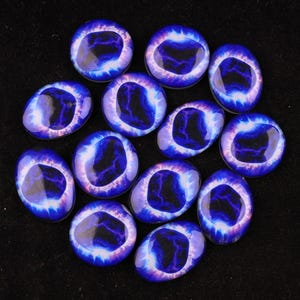 Pode incluir: Uma coleção de doze cabochões de vidro em forma de olho. Cada cabochão apresenta um design de olho detalhado com uma íris azul, uma pupila preta e um anel externo branco e roxo. Os cabochões estão dispostos sobre uma superfície preta.