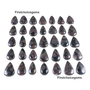 Może przedstawiać: Kolekcja kamieni szlachetnych w kształcie łezki z mieszanką ciemnoczerwonych i zielonych odcieni. Kamienie szlachetne ułożone są na białej powierzchni, z tekstem "Firstchoicegems" u góry i u dołu.