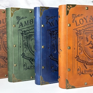 May include: Four leather-bound journals in shades of brown, green, blue, and orange. Each journal features decorative corner accents, metal studs, and embossed text and imagery. The orange journal displays the word "Loyalty" and a crest.