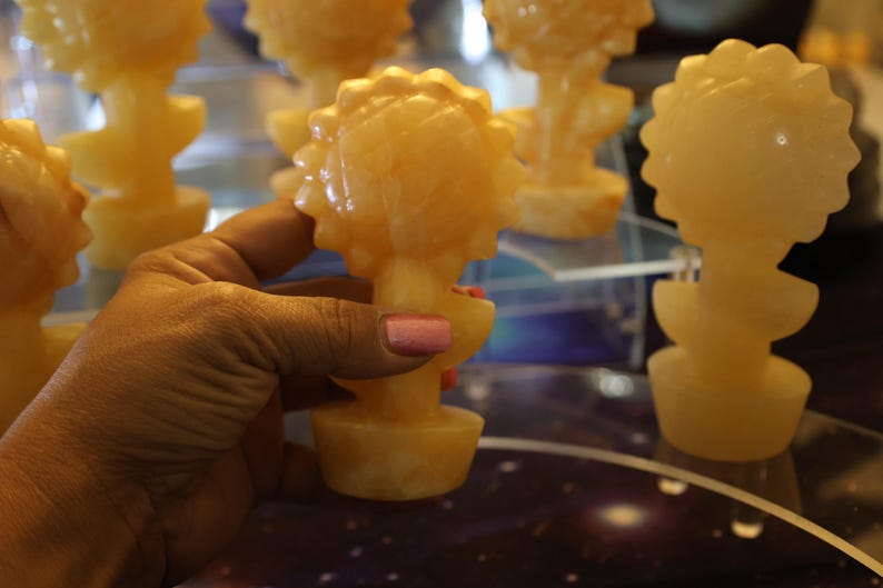 May include: A collection of five small, hand-carved, yellow-orange, flower-shaped sculptures. The sculptures are made of a translucent material, possibly alabaster or calcite. The sculptures are arranged on a table with a dark, starry surface.