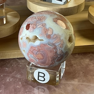 May include: A spherical stone featuring a mix of cream, dusty pink, and deep red patterns with small crystalline cavities. The sphere rests on a clear square acrylic stand labeled with a white circular sticker containing the letter B.
