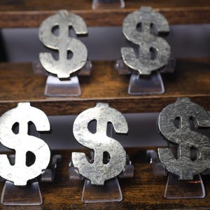 May include: Six metallic dollar sign sculptures displayed on a wooden shelf. The dollar signs are silver-colored and mounted on clear plastic stands. The shelf is made of dark brown wood.