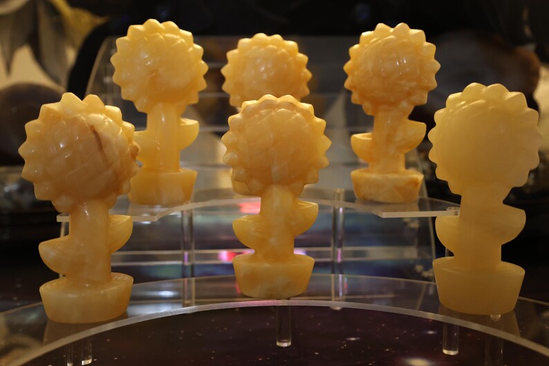 May include: Six small, hand-carved, yellow-orange, alabaster sculptures of sunflowers. Each sculpture is about 4 inches tall and features a detailed sunflower head and stem.