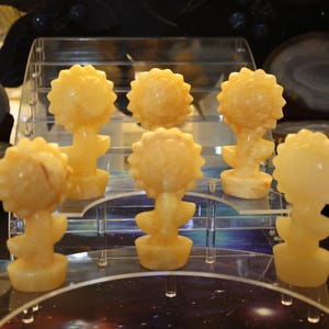 May include: Six yellow carved sunflower sculptures on a clear acrylic display stand. The sculptures are made of polished stone and have a smooth, glossy finish.