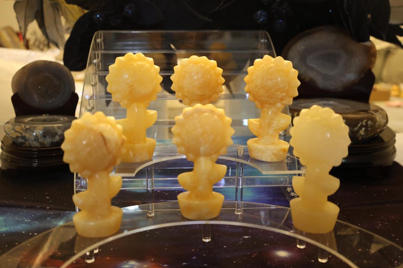 May include: Six yellow carved sunflower sculptures on a clear acrylic display stand. The sculptures are made of polished stone and have a smooth, glossy finish.