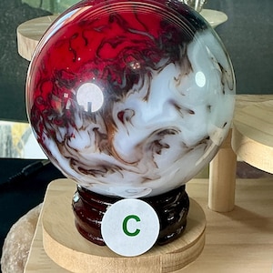 May include: A spherical glass orb featuring a swirling pattern of deep red, opaque white, and dark brown colors. The orb is mounted on a dark brown, circular wooden stand and features a small white circular sticker with the green letter C.