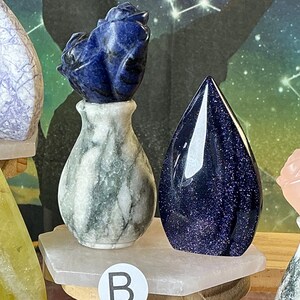 May include: A small white and grey marble-like vase containing a carved dark blue sodalite rose, positioned next to a smooth, teardrop-shaped dark blue goldstone crystal with glittery inclusions, both resting on a white hexagonal stone base labeled B.