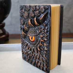 May include: A black leather-bound journal with a detailed embossed dragon design. The dragon's eye is a bright orange color.