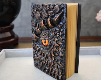 May include: A black leather-bound journal with a detailed embossed dragon design. The dragon's eye is a bright orange color.