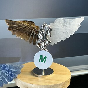 May include: A silver-toned metal figurine of two figures embracing, featuring a brown tiger's eye wing on the left and a clear white quartz wing on the right. A white circular tag with the green letter M is attached to the base of the figurine.