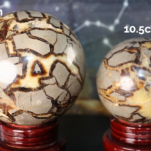 May include: Two decorative spheres, 13cm and 10.5cm in diameter, rest on red wooden stands. The spheres have a light gray base color with brown and tan veining patterns. The spheres are polished and have a glossy finish.
