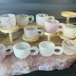 May include: A collection of small, translucent cups carved from light pink and white quartz stone. Each cup features a rounded bowl and a circular handle, displaying natural crystalline textures and variations in opacity across the set.