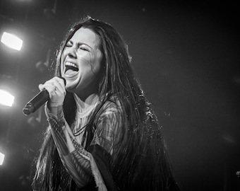 May include: A woman with long dark hair sings into a microphone while performing on stage. The stage is lit with bright lights.