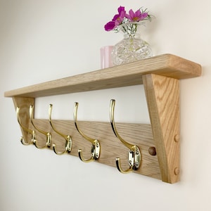 May include: A wooden shelf with five brass hooks, mounted on a white wall. The shelf has a light wood finish and a slightly angled design. A small pink vase with pink flowers sits on the shelf.