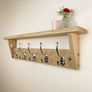 May include: A wooden shelf with six chrome hooks, mounted on a wall. The shelf has a light wood finish and is decorated with a small vase of red flowers and a pink candle.