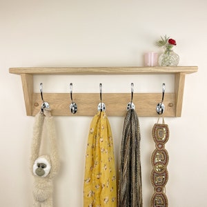 May include: A wooden coat rack with five hooks and a shelf. The rack is made of light wood and has a natural finish. There are three items hanging on the hooks: a yellow scarf, a brown scarf, and a gold beaded belt.