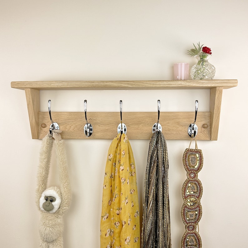 May include: A wooden coat rack with five hooks and a shelf. The rack is made of light wood and has a natural finish. There are three items hanging on the hooks: a yellow scarf, a brown scarf, and a gold beaded belt.