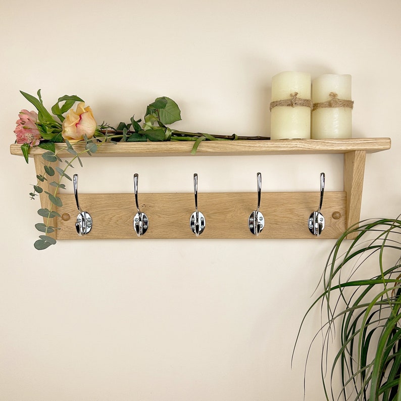May include: A wooden shelf with five chrome hooks, a shelf for storage, and two white candles. The shelf is mounted on a white wall.