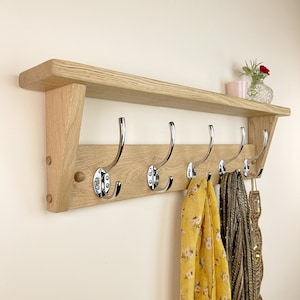 May include: A wooden coat rack with a shelf and five chrome hooks. The rack is mounted on a wall and has a light wood finish. There are scarves hanging on the hooks.