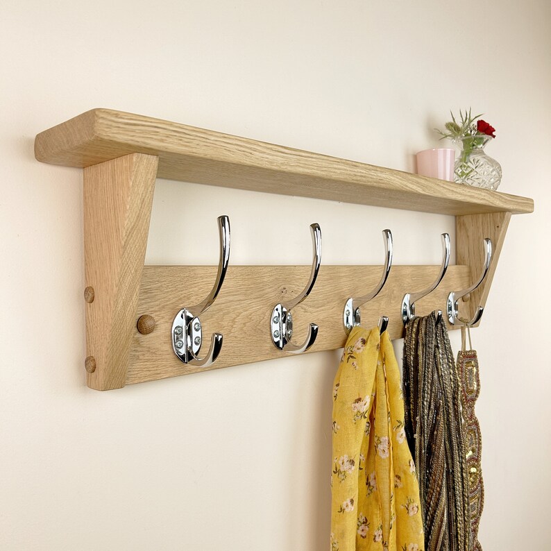 May include: A wooden coat rack with a shelf and five chrome hooks. The rack is mounted on a wall and has a light wood finish. There are scarves hanging on the hooks.