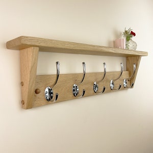 May include: A wooden wall-mounted shelf with five chrome hooks. The shelf is made of natural oak wood and has a slightly angled design.