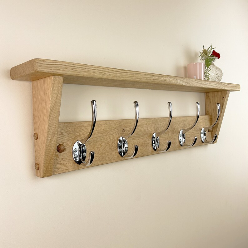 May include: A wooden wall-mounted shelf with five chrome hooks. The shelf is made of natural oak wood and has a slightly angled design.