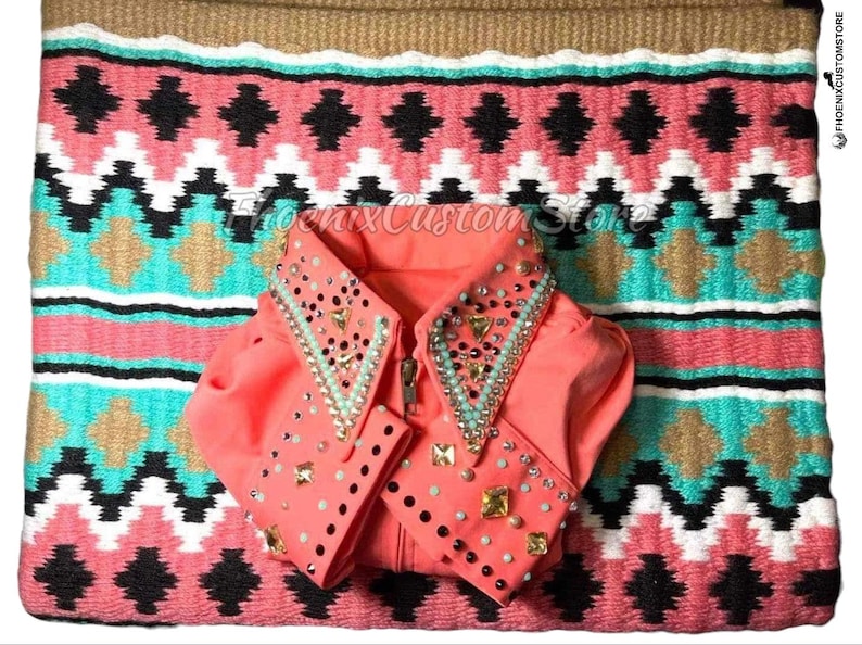 May include: A coral-colored equestrian show shirt with a decorative collar and cuffs, adorned with sparkling embellishments. The shirt is displayed on a patterned saddle blanket with a geometric design in coral, turquoise, black, and gold.