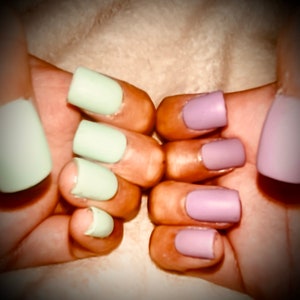 May include: Close-up of hands with light green and lavender nail polish. The nails are short and rounded.