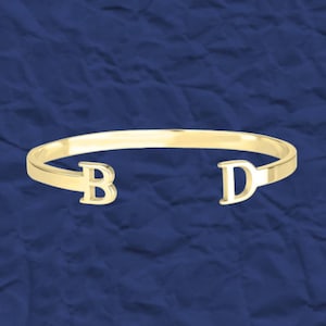 May include: A gold bangle bracelet with the letters "B" and "D" on either side.