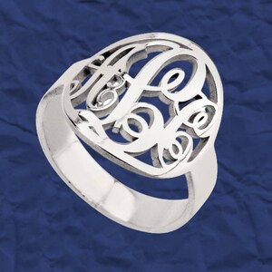 K&ouml;nnte beinhalten: Ein silberner Ring mit einem Monogramm-Design mit drei ineinander verschlungenen Buchstaben. Der Ring ist aus glattem, poliertem Metall gefertigt und hat ein schlichtes Band.