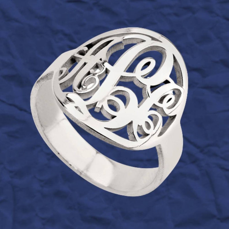K&ouml;nnte beinhalten: Ein silberner Ring mit einem Monogramm-Design mit drei ineinander verschlungenen Buchstaben. Der Ring ist aus glattem, poliertem Metall gefertigt und hat ein schlichtes Band.