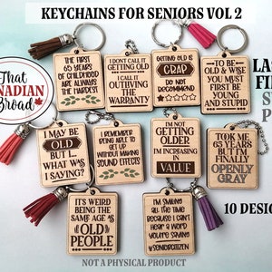 May include: Ten laser cut wooden keychains with humorous sayings about getting older. The keychains are brown with white text and are attached to silver chains with tassels. The text on the keychains includes phrases like "I'm not getting older, I'm increasing in value" and "Took me 65 years, but I'm finally openly grey".
