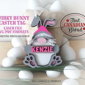 Puede incluir: Una etiqueta de Pascua de gnomo conejito rosa y gris con el nombre "Kenzie" en ella. La etiqueta est&aacute; rodeada de huevos de Pascua blancos y hierba verde. El texto "That Canadian Broad" es visible en la esquina superior derecha de la imagen.