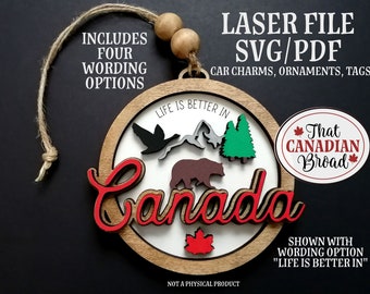 Op de afbeelding: Een ronde houten ornament met het woord "Canada" in rode letters. De ornament heeft een beer, bomen, bergen en een vogel. De tekst "Life is better in" staat rond de bovenkant van de ornament. De tekst "That Canadian Broad" staat in een cirkel aan de rechterkant van de ornament. De ornament is afgebeeld met een touwtje om op te hangen.