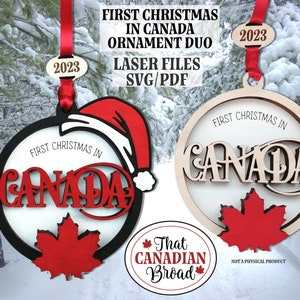 Könnte beinhalten: Zwei lasergeschnittene Weihnachtsbaumschmuck-Designs aus Holz mit den Worten "First Christmas in Canada" und einem roten Ahornblatt. Die Ornamente befinden sich in einer verschneiten Winterlandschaft mit einem roten Band und einem Etikett mit der Aufschrift "2023".