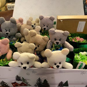 May include: A collection of handmade teddy bears in a decorative box. The bears are in various colors, including pink, gray, and cream, with black button eyes and stitched features. The box is decorated with a winter scene.