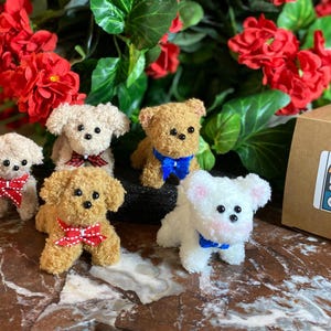May include: Five plush toy dogs with bows, in cream, tan, and white, are arranged in front of red flowers and green leaves. A brown box with a sticker that says "DKMS Crafts" is on the right.