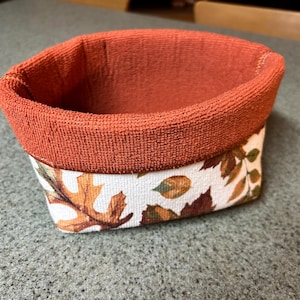 May include: A small fabric basket with a textured, solid burnt orange rim and a white exterior featuring a printed pattern of various autumn leaves in shades of brown, orange, and green. The basket has a rounded rectangular shape and open top.