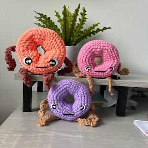 May include: Three handmade crochet donuts in shades of peach, pink, and purple, each with a hole in the center. They have black eyes, stitched mouths, and brown crocheted legs. The donuts are arranged on a gray surface.