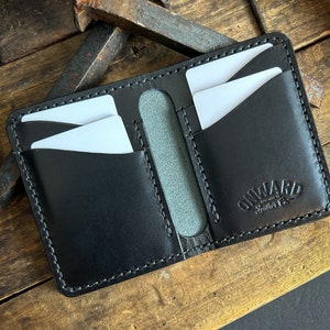 Op de afbeelding: Zwart leren portemonnee met witte stiksels en meerdere kaartsleuven. De portemonnee heeft het logo "ONWARD Leather Co.". Er zijn verschillende witte kaarten zichtbaar. De portemonnee ligt op een rustiek houten oppervlak.