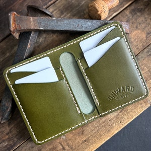 Op de afbeelding: Olijfgroene leren portemonnee met witte stiksels en meerdere kaartsleuven. De portemonnee heeft de tekst "ONWARD Leather Co." in reliëf op de voorkant. De portemonnee is open en onthult witte kaarten binnenin. De achtergrond omvat een rustiek houten oppervlak en een metalen object.