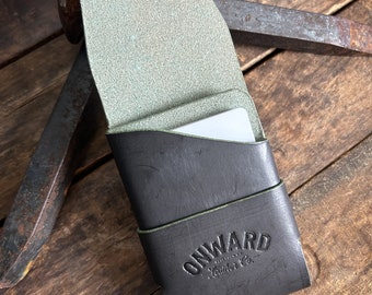 Peut inclure: Porte-cartes en cuir noir avec intérieur vert clair. Le porte-cartes porte l'inscription "ONWARD Leather Co." en relief sur le devant. Le porte-cartes est ouvert, révélant une carte blanche à l'intérieur. Fond en bois avec outils métalliques.