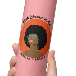 May include: Pink stainless steel tumbler with a black and orange circle graphic. The graphic features a black woman with an afro, sunglasses, and earrings. The text "Black Behavior Analyst shaping my fro & behavior" is printed around the graphic.
